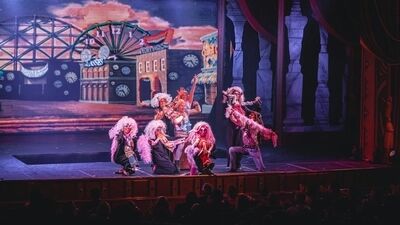 Een groep performers in kleurrijke kostuums voert een scène op in een theater met een levendige achtergrond.